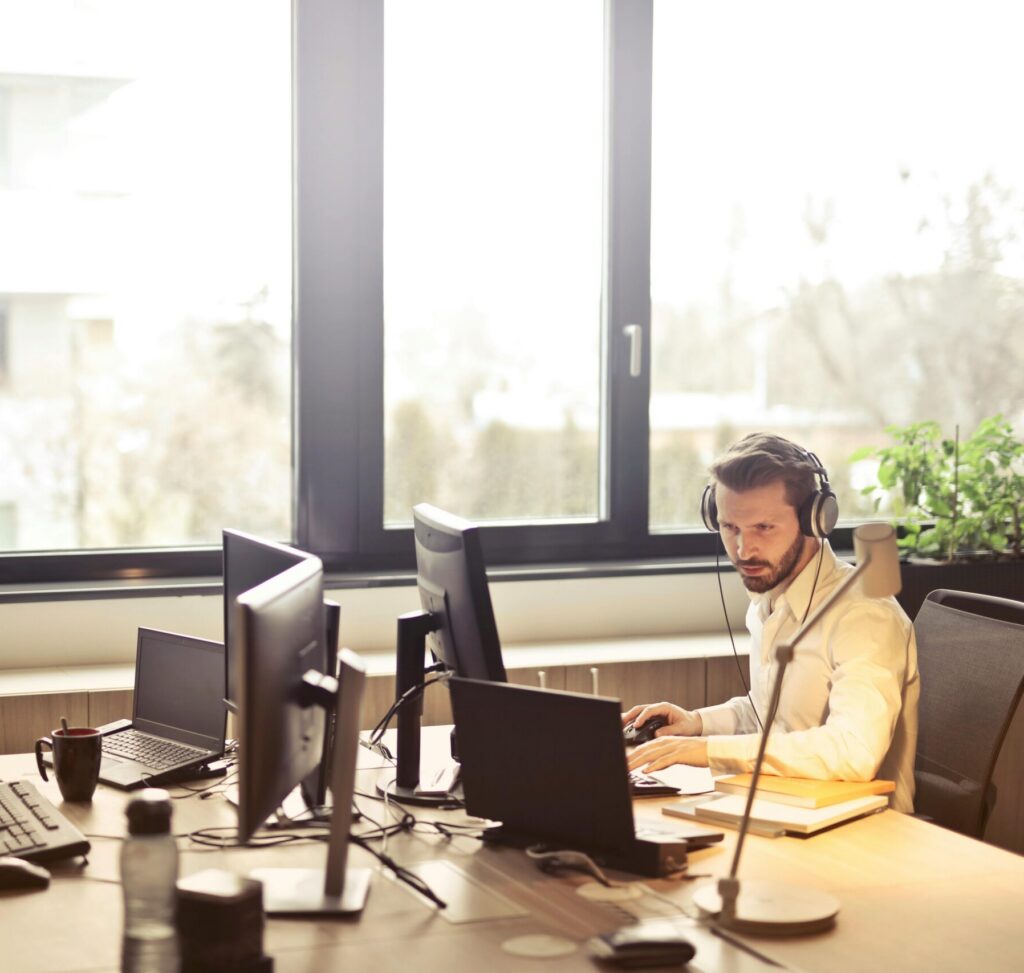 A businessman sits at a desk using multiple computers and a headset in a well-lit modern office.